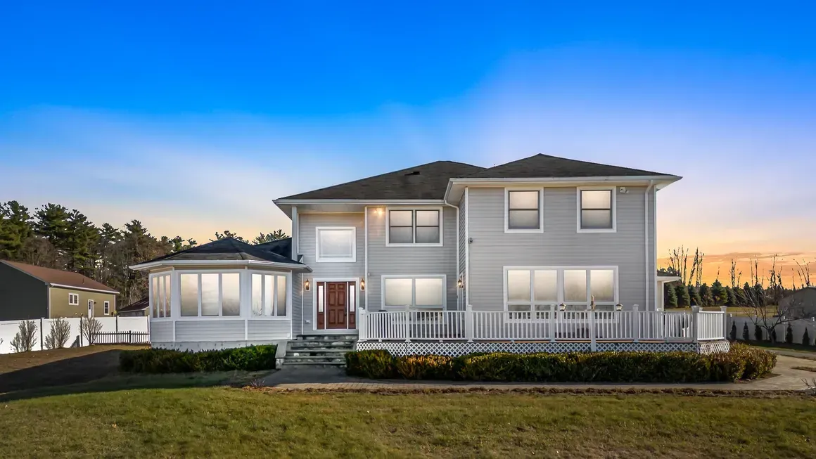 Two-story gray house with a porch and deck on a grassy lot under a blue sky.