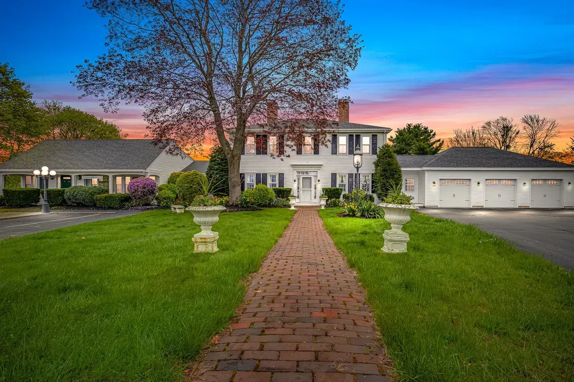 A brick pathway leads to a white colonial-style house with a three-car garage; green lawn and sunset sky.