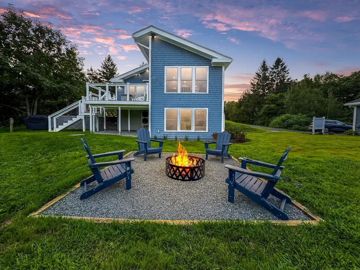 Blue house with fire pit, Adirondack chairs, and lake view at sunset.