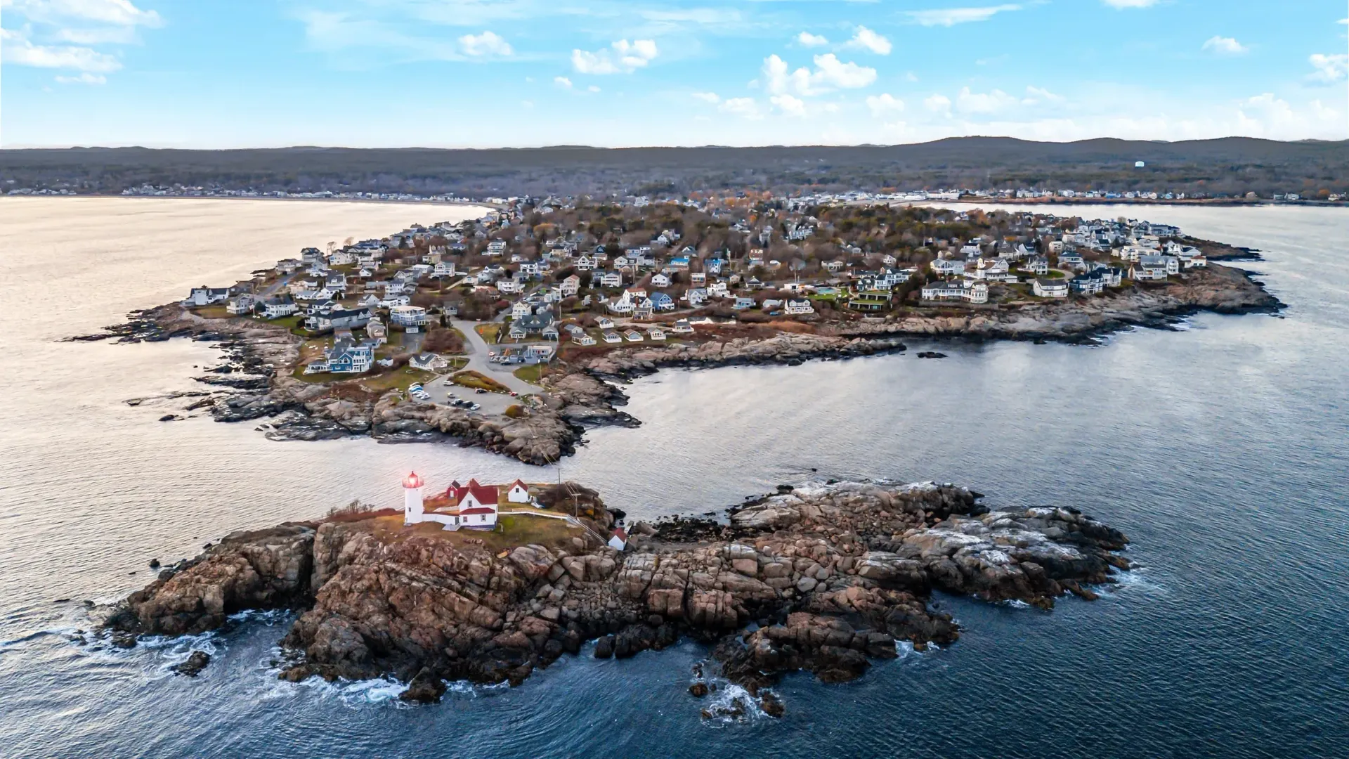 Aerial view of a coastal island with a lighthouse on a rocky outcrop and a town with houses.