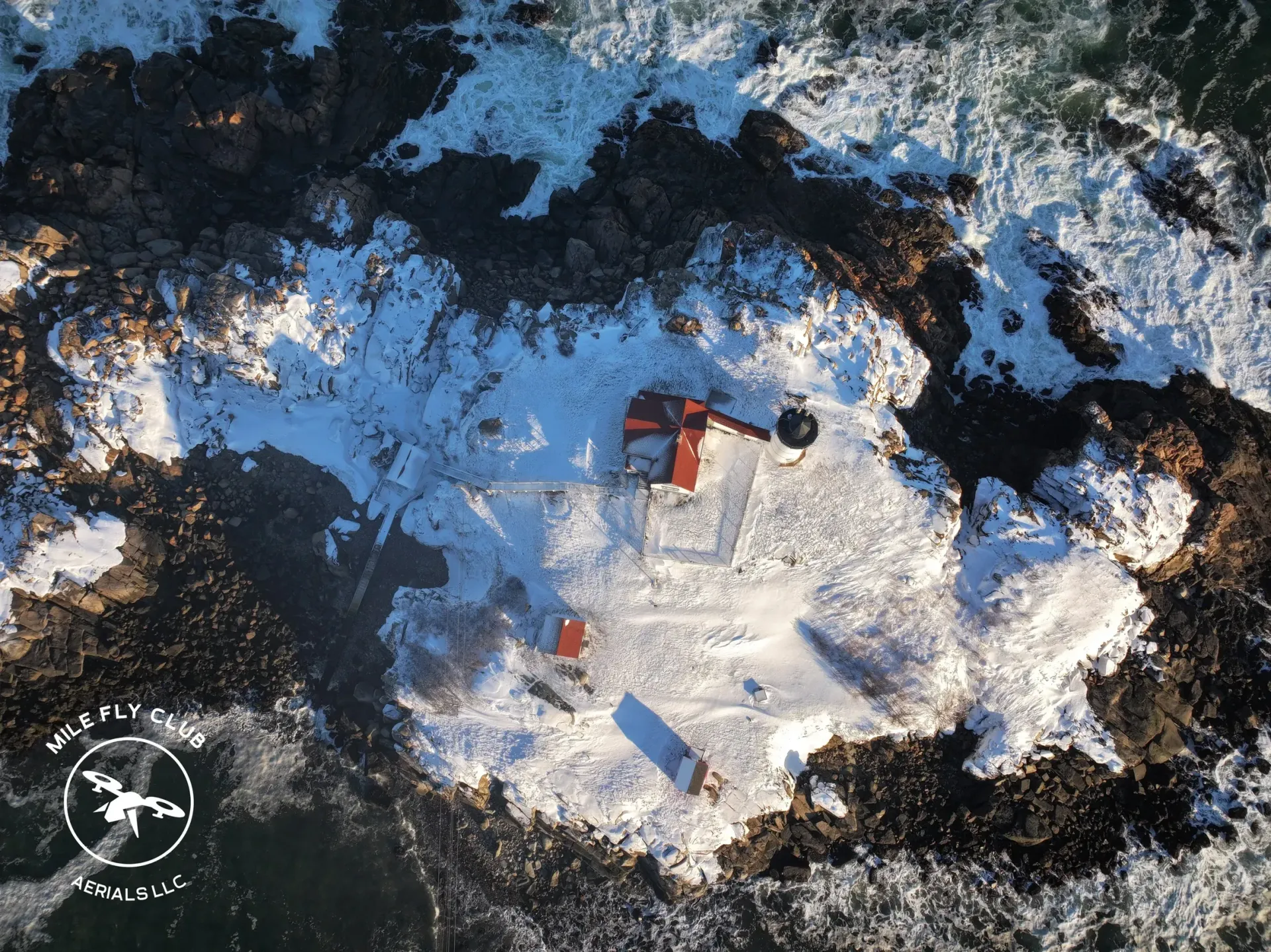 Aerial view of a snowy island with a red lighthouse. Ocean surrounds the rocky island.