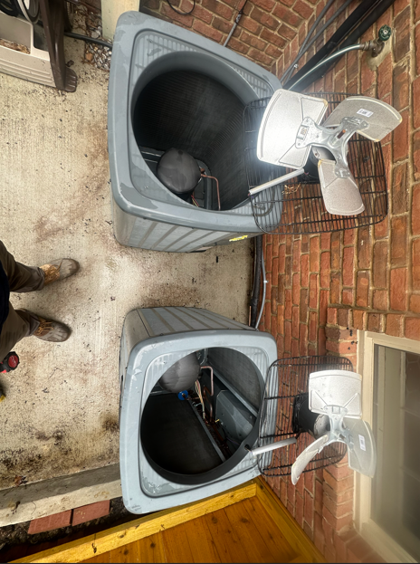 Two gray air conditioning units next to a brick wall. A person's legs and shoes are in the frame.