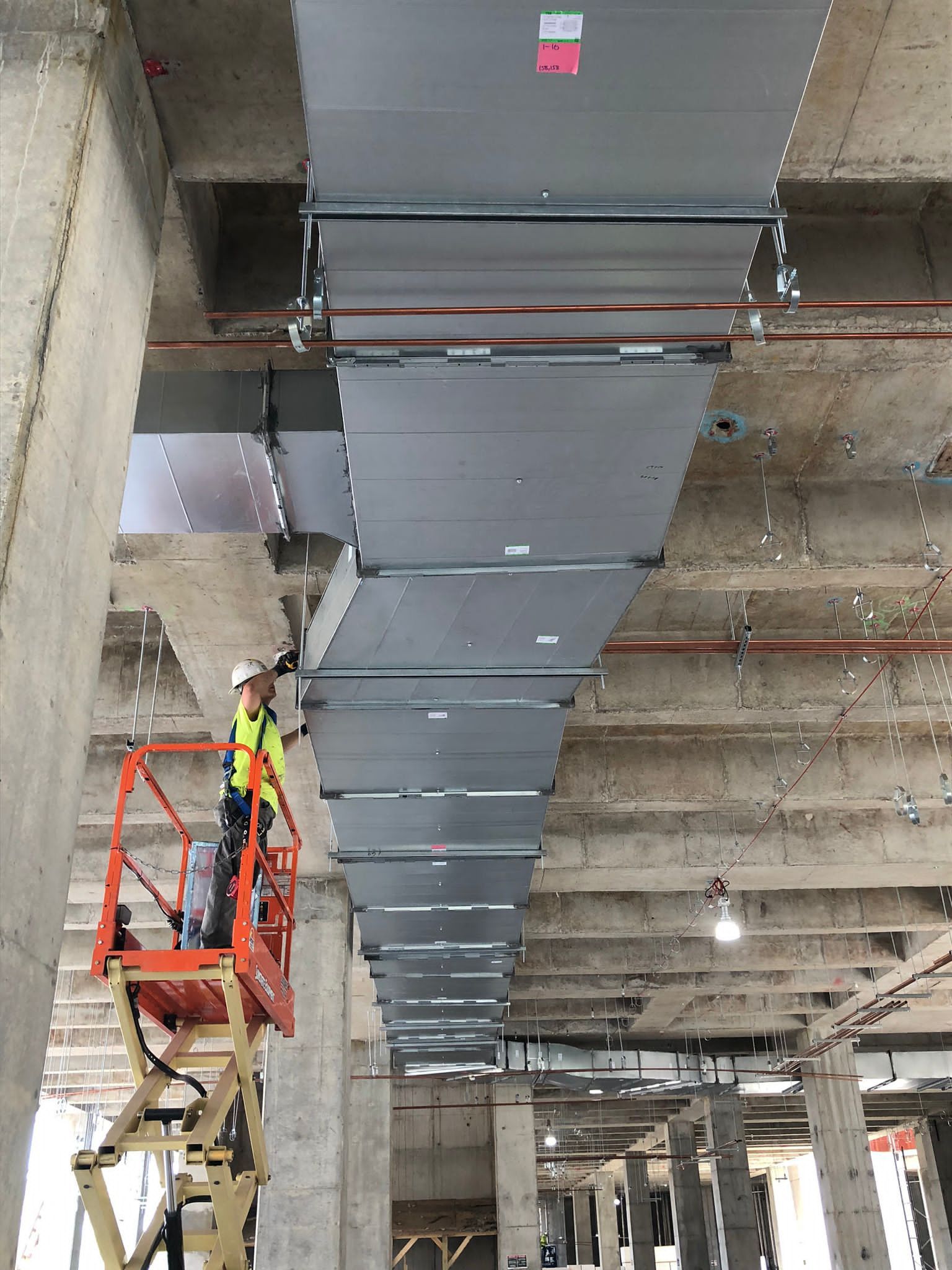 Person on lift installs rectangular ductwork in a concrete building.
