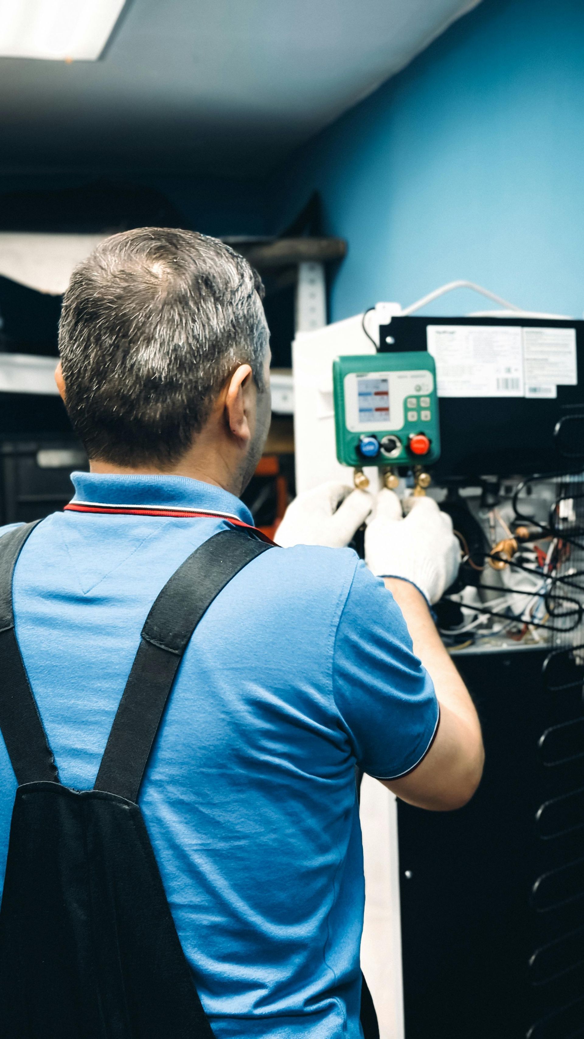 Technician in blue shirt and overalls works on equipment, wearing gloves, in a shop setting.