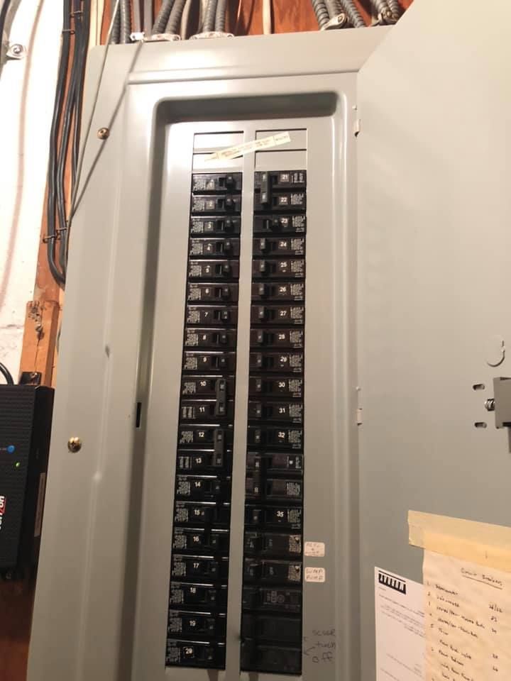 Electrical panel with numerous circuit breakers, housed in a gray metal enclosure. Door is open.
