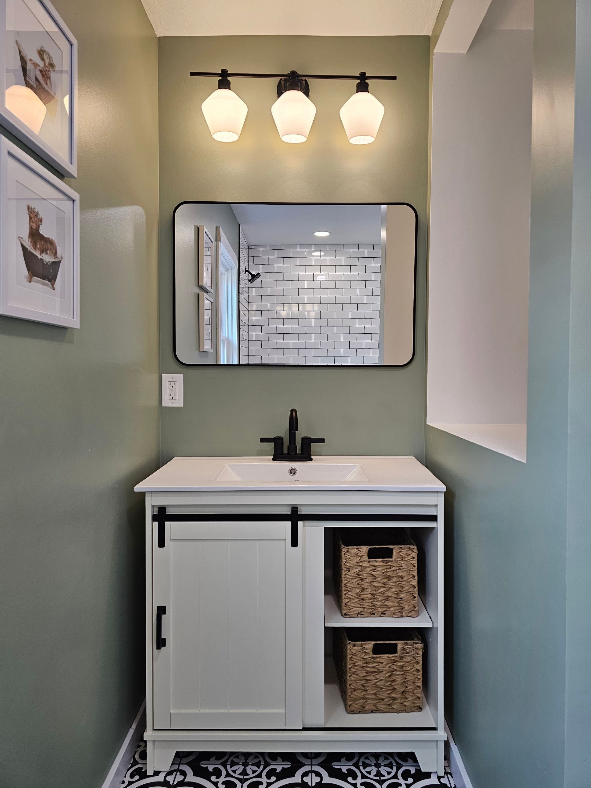 Small bathroom with white vanity, black faucet, and green walls. Mirror and three-bulb light fixture above.