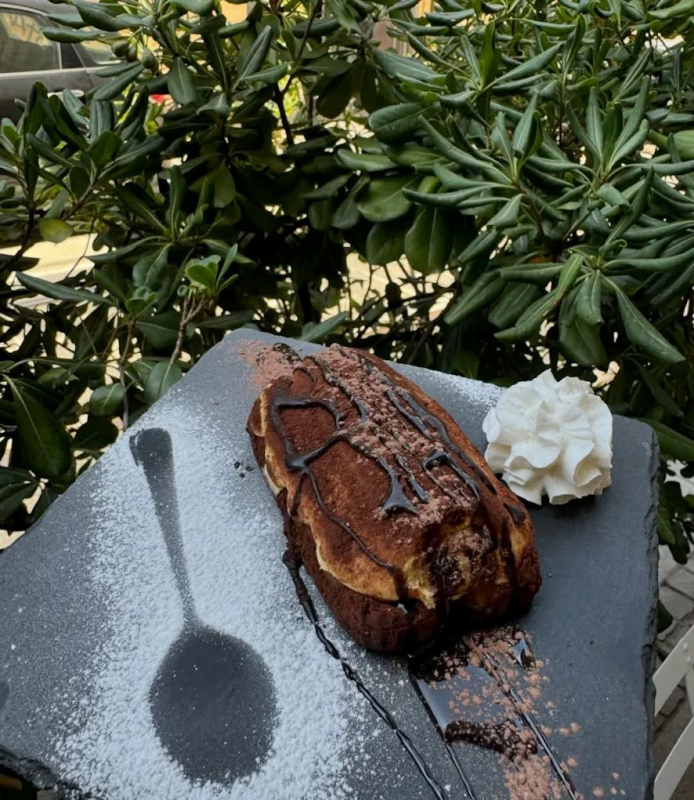 Dessert tiramisù servito su una lastra di pietra scura, spolverato con cacao in polvere e guarnito con panna montata e salsa al cioccolato.