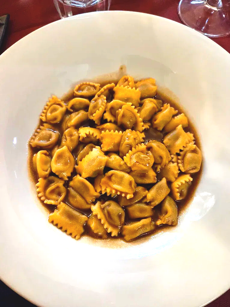 Piatto di ravioli in salsa scura. I ravioli sono dorati, la salsa è marrone, su un piatto bianco.