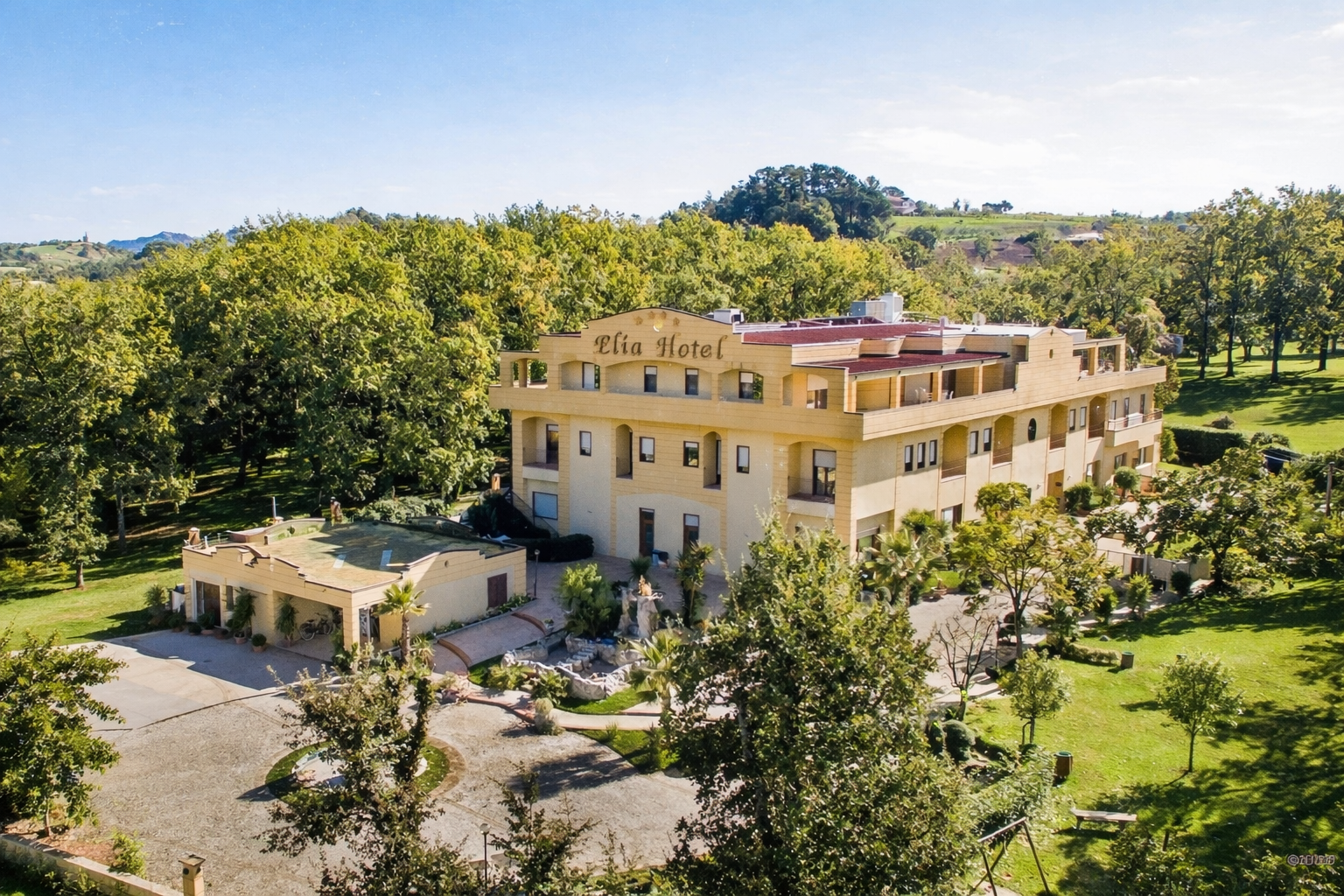 Vista aerea di un edificio alberghiero color crema circondato da alberi e vegetazione in una giornata di sole.