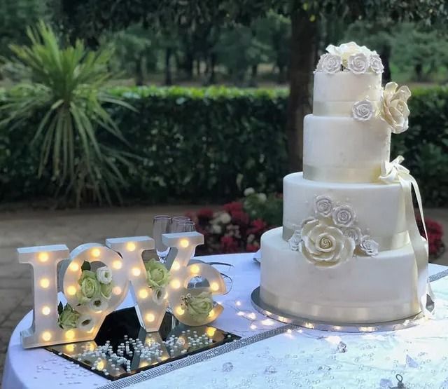 Una torta nuziale bianca a quattro piani con decorazioni floreali è posta accanto a un cartello luminoso