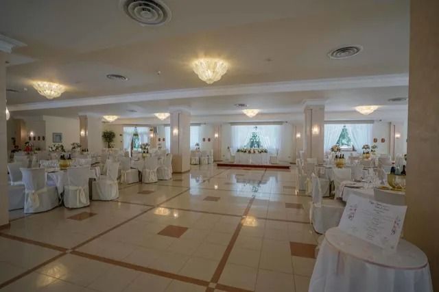 Ampia sala per banchetti ben illuminata, con file di tavoli da pranzo rotondi coperti da tovaglie e sedie bianche.