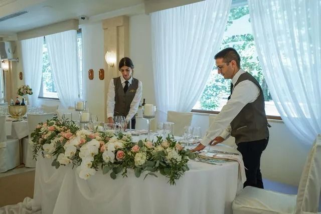In una luminosa ed elegante sala per eventi, i camerieri in gilet apparecchiano un tavolo rotondo
