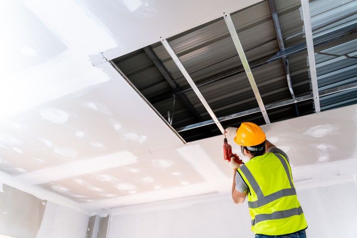 Construction worker installing ceiling drywall, wearing a yellow hardhat and safety vest.