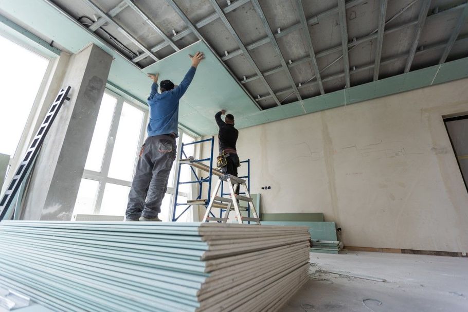 Two workers install drywall ceiling in a room under construction; drywall sheets stacked below.