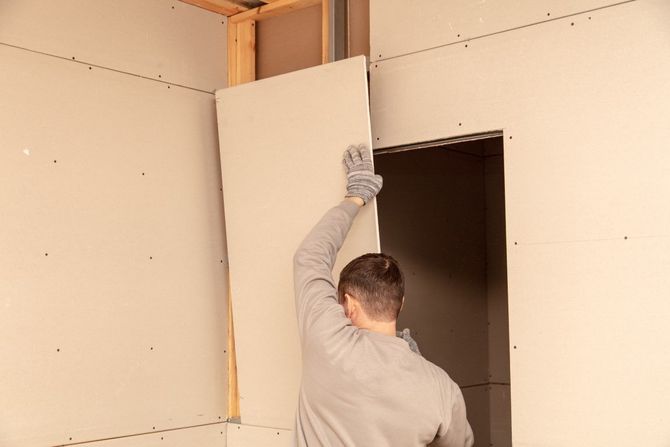 Person installing drywall in a room with an unfinished doorway; wearing gloves.
