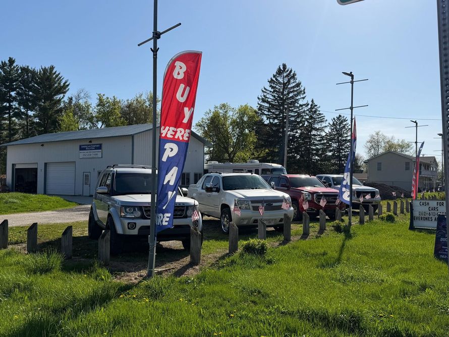 Used car lot with parked trucks and SUVs beside a red-and-blue feather flag outdoors
