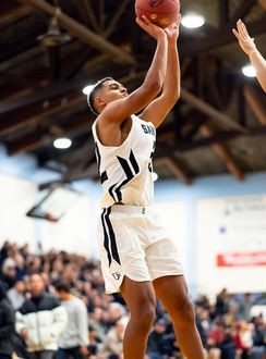 Basketball player in a white uniform with black accents shoots a basketball. He's in a gymnasium with a crowd in the background.