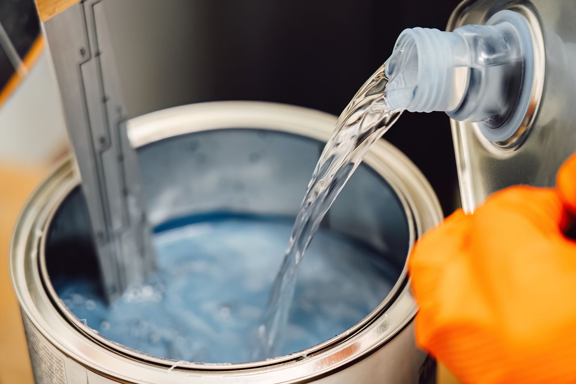 Orange-gloved hand pouring liquid into a metal bucket from a spout