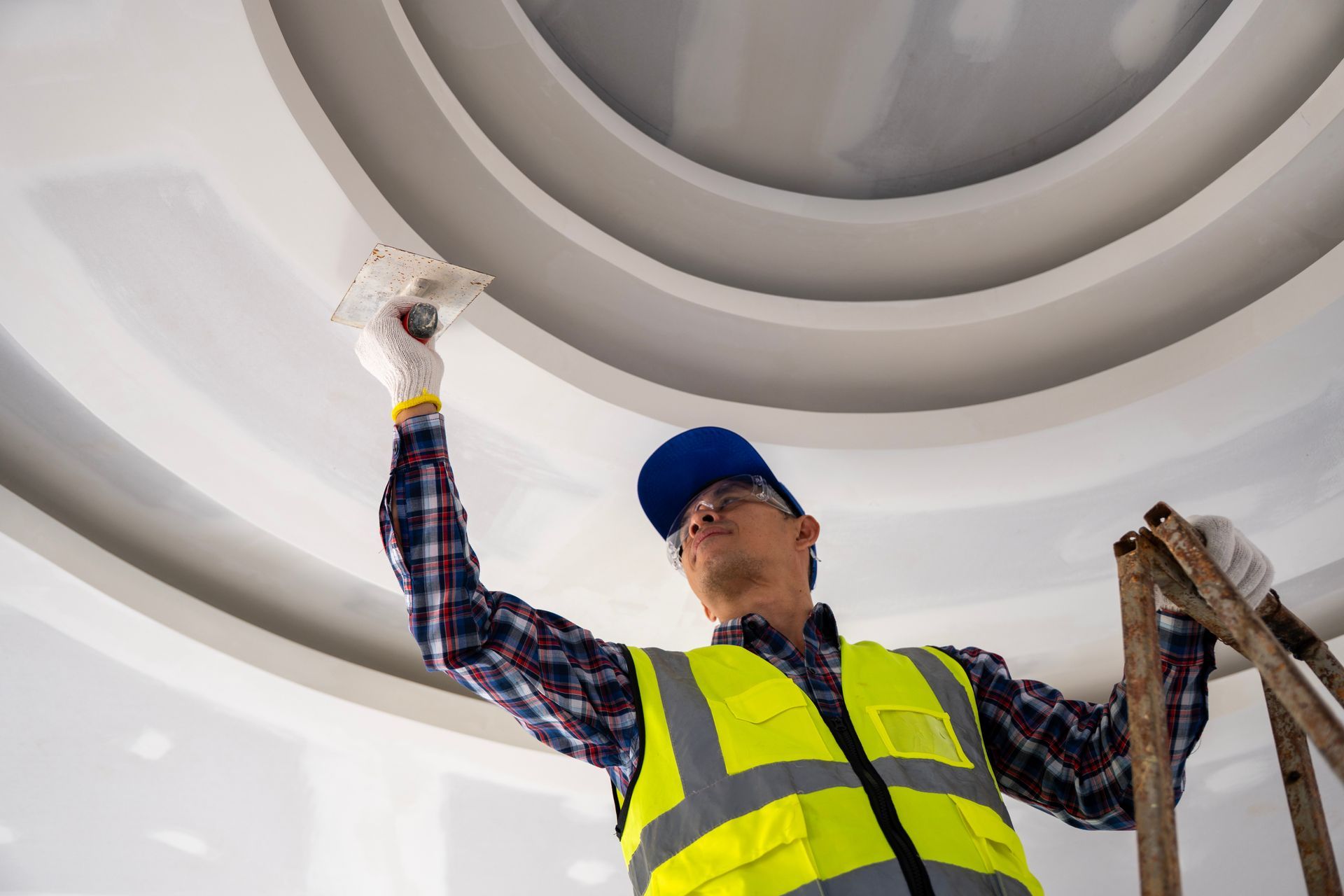 Worker in a safety vest painting a white ceiling near a ladder