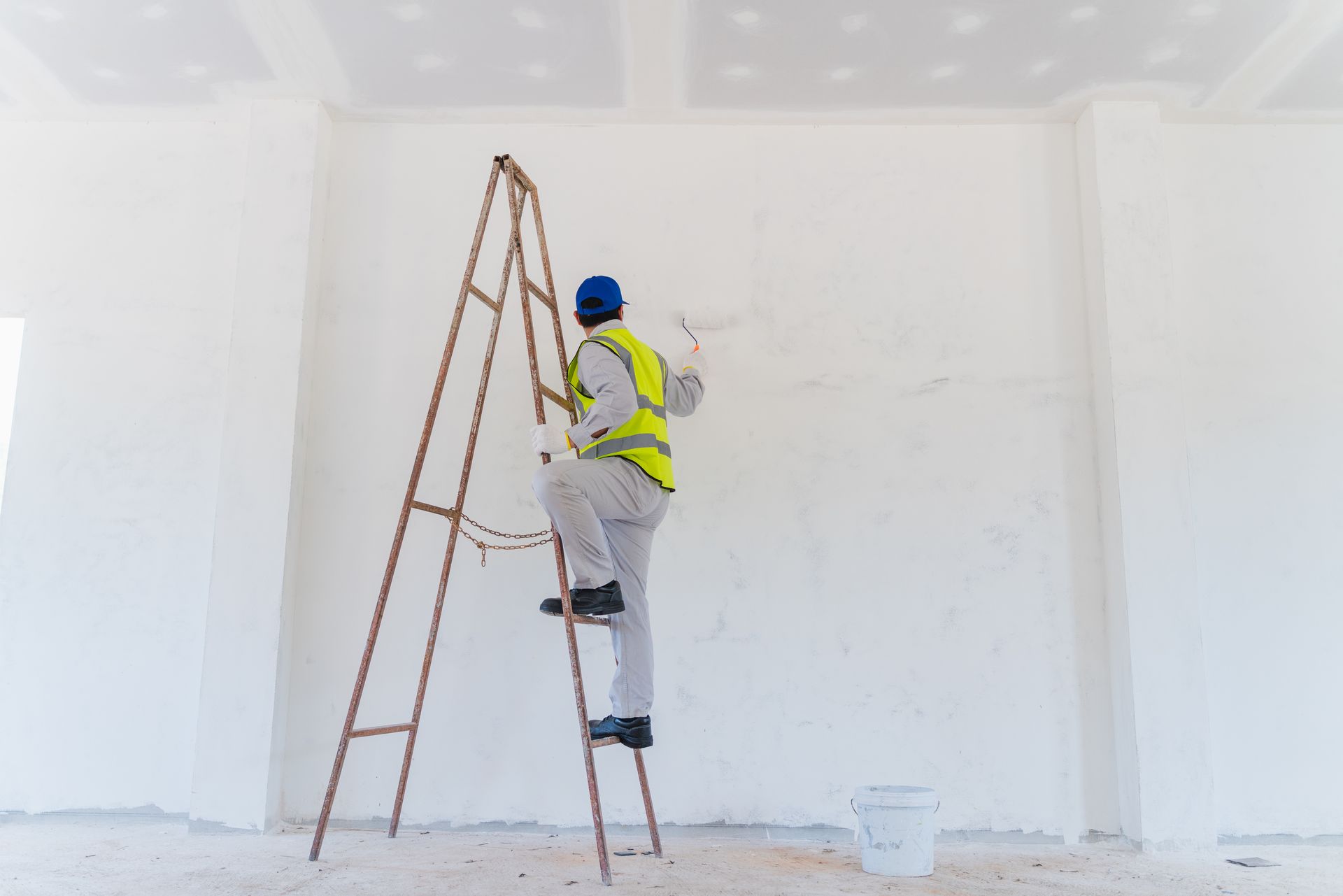Worker on a ladder painting a white room in a blue helmet and yellow safety vest