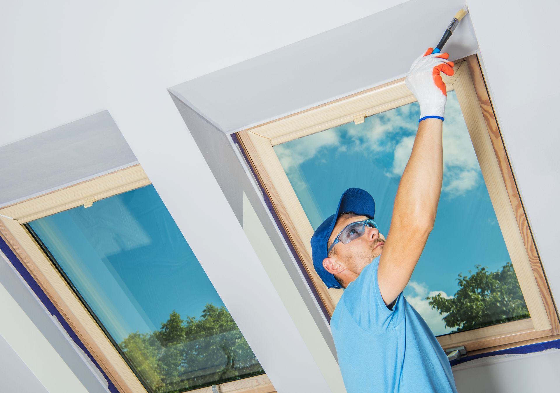 Worker painting a window frame while standing on a ladder, with blue sky and trees reflected in the glass