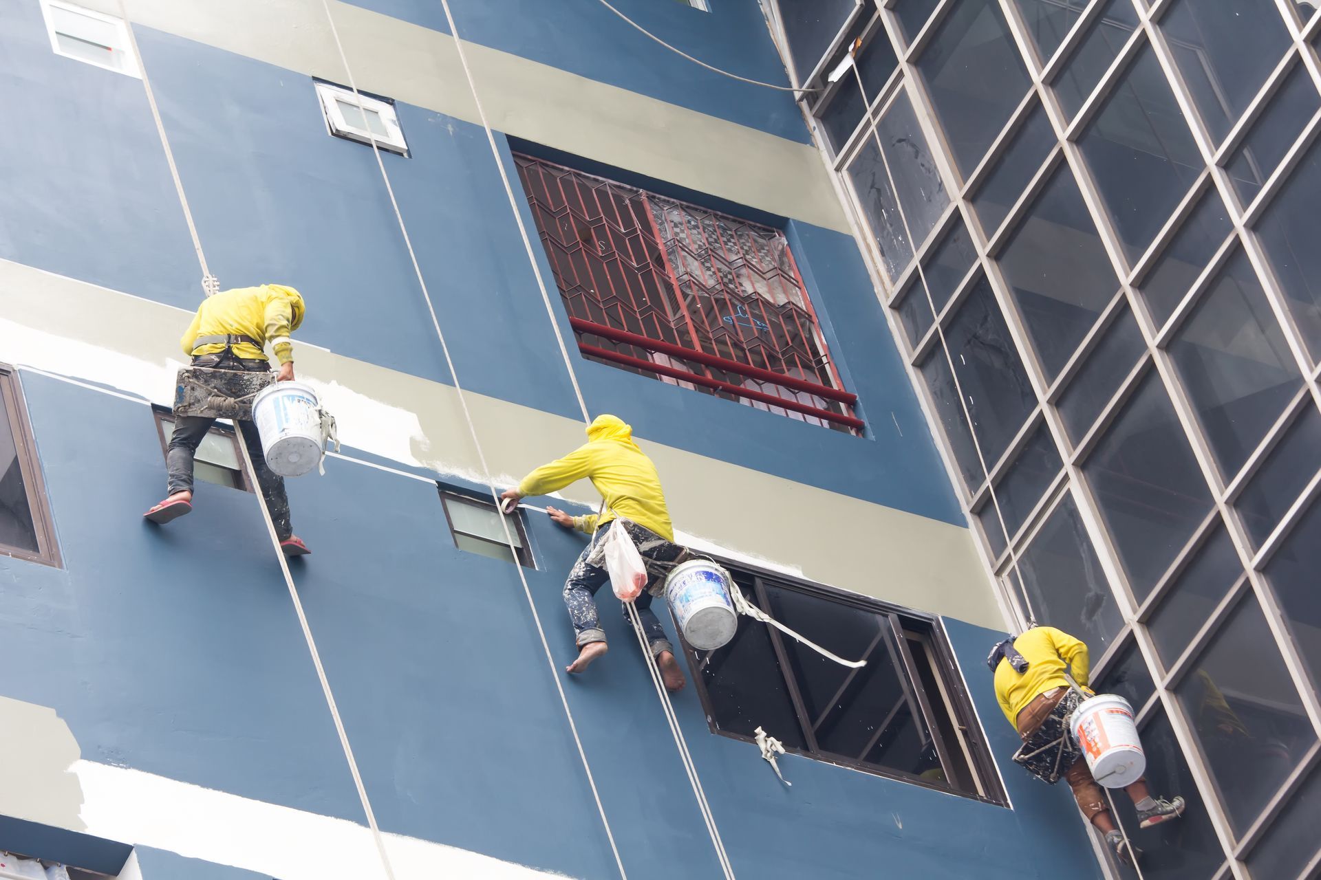 Three window cleaners in yellow jackets rappel down a blue high-rise building.