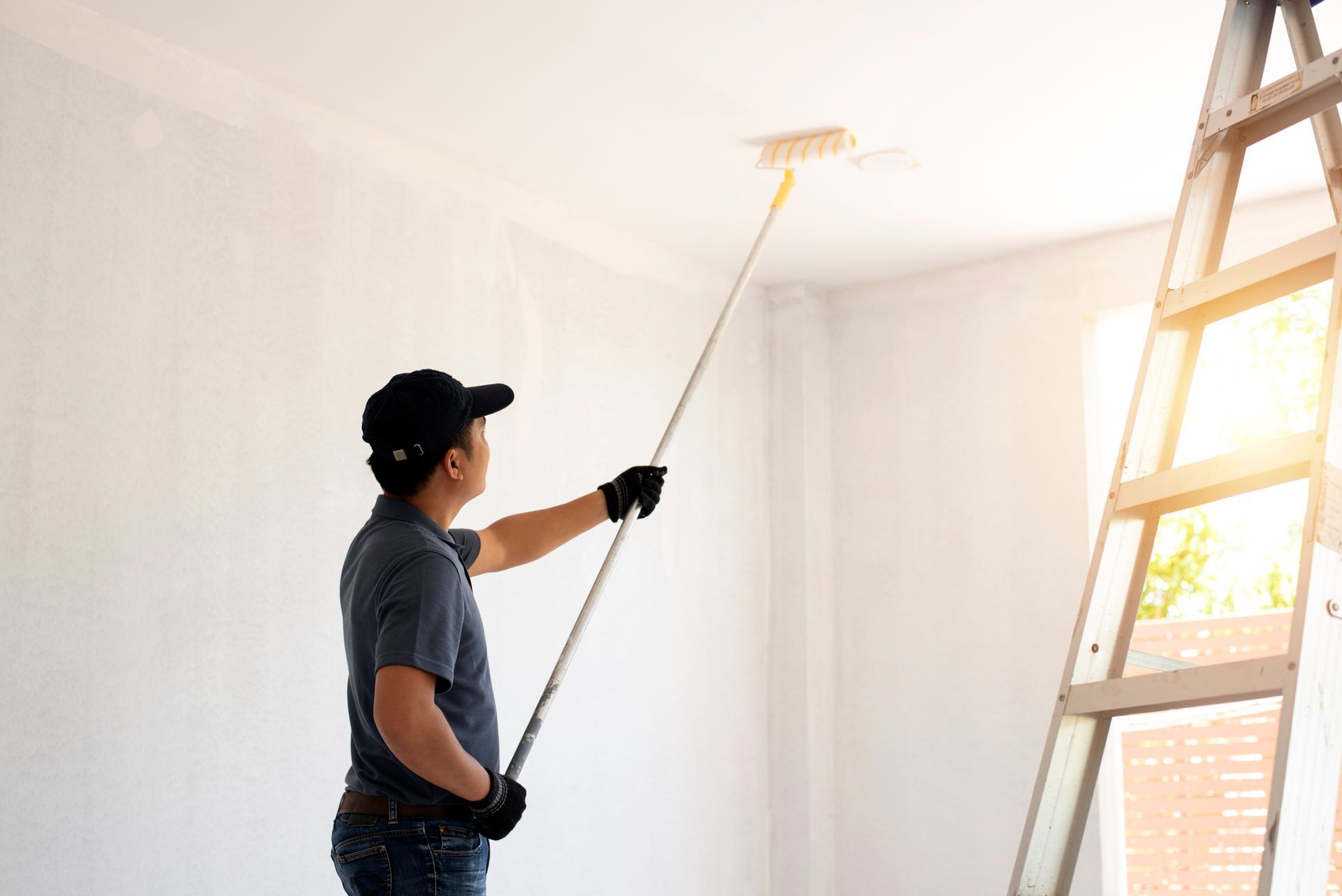Person painting a white ceiling beside a ladder in a bright room