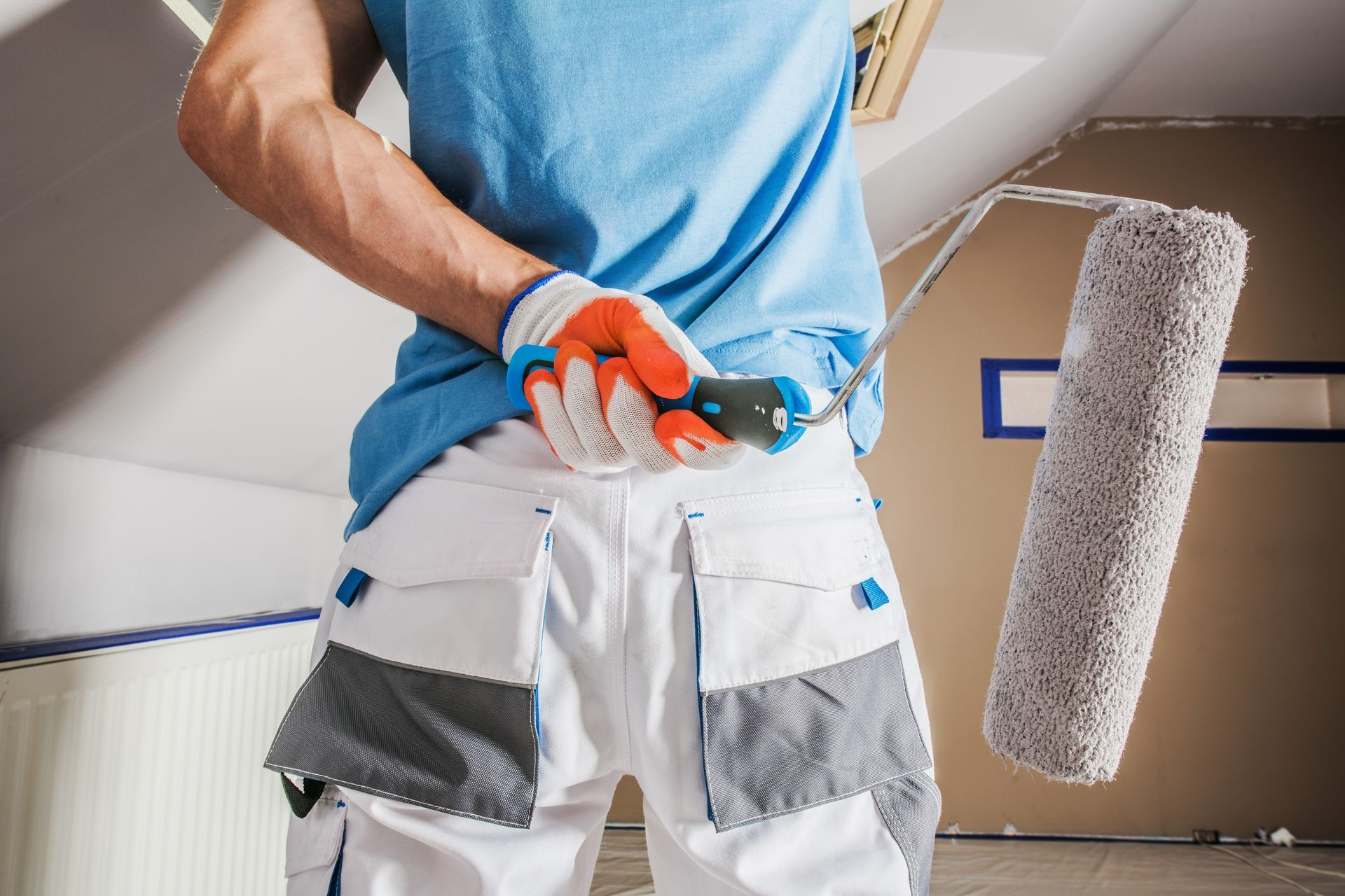 Painter using a roller and paint sprayer in a room with white walls and gray floor-end trim.