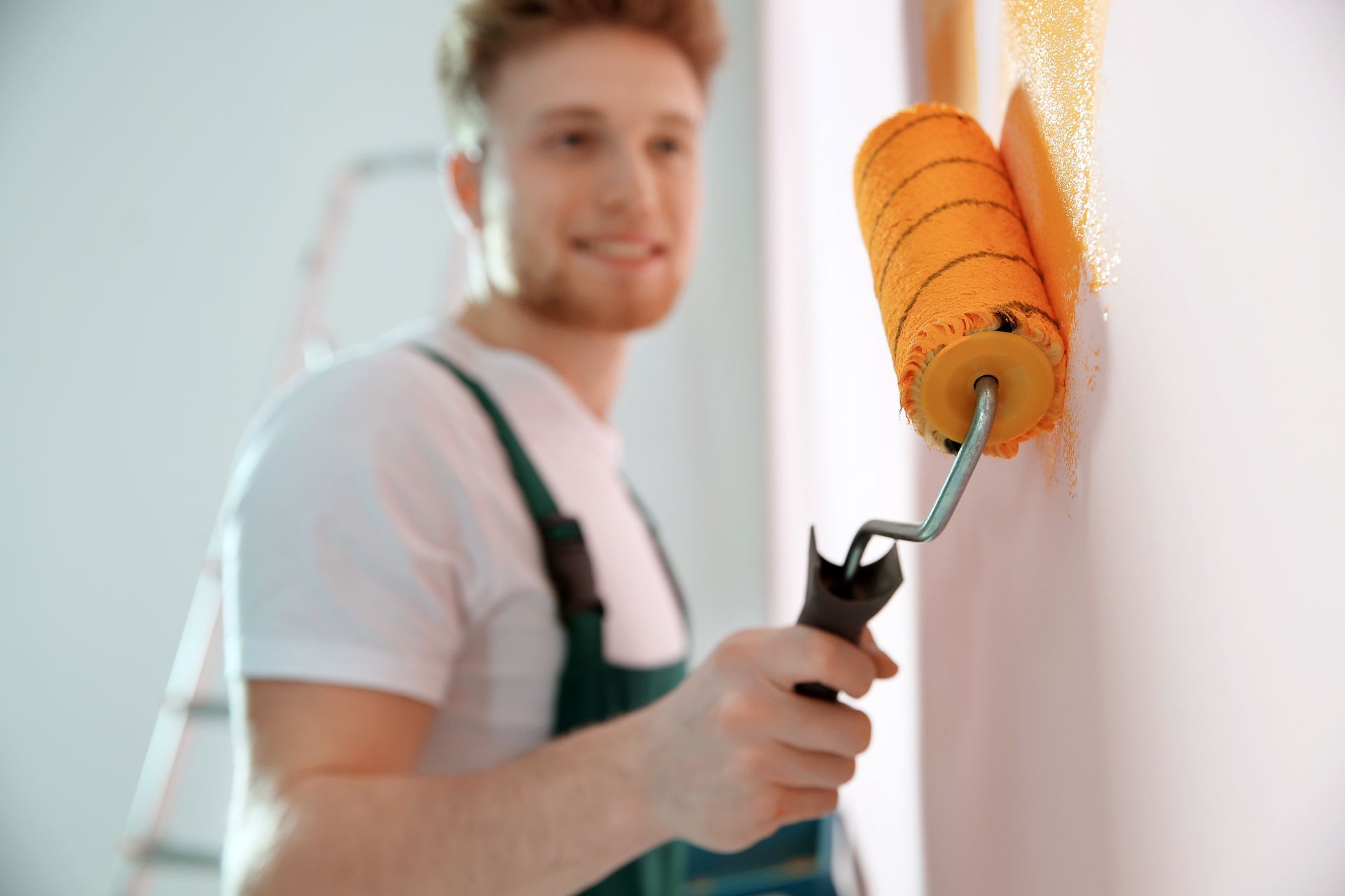 Person painting a wall with an orange roller, wearing white shirt and green overalls