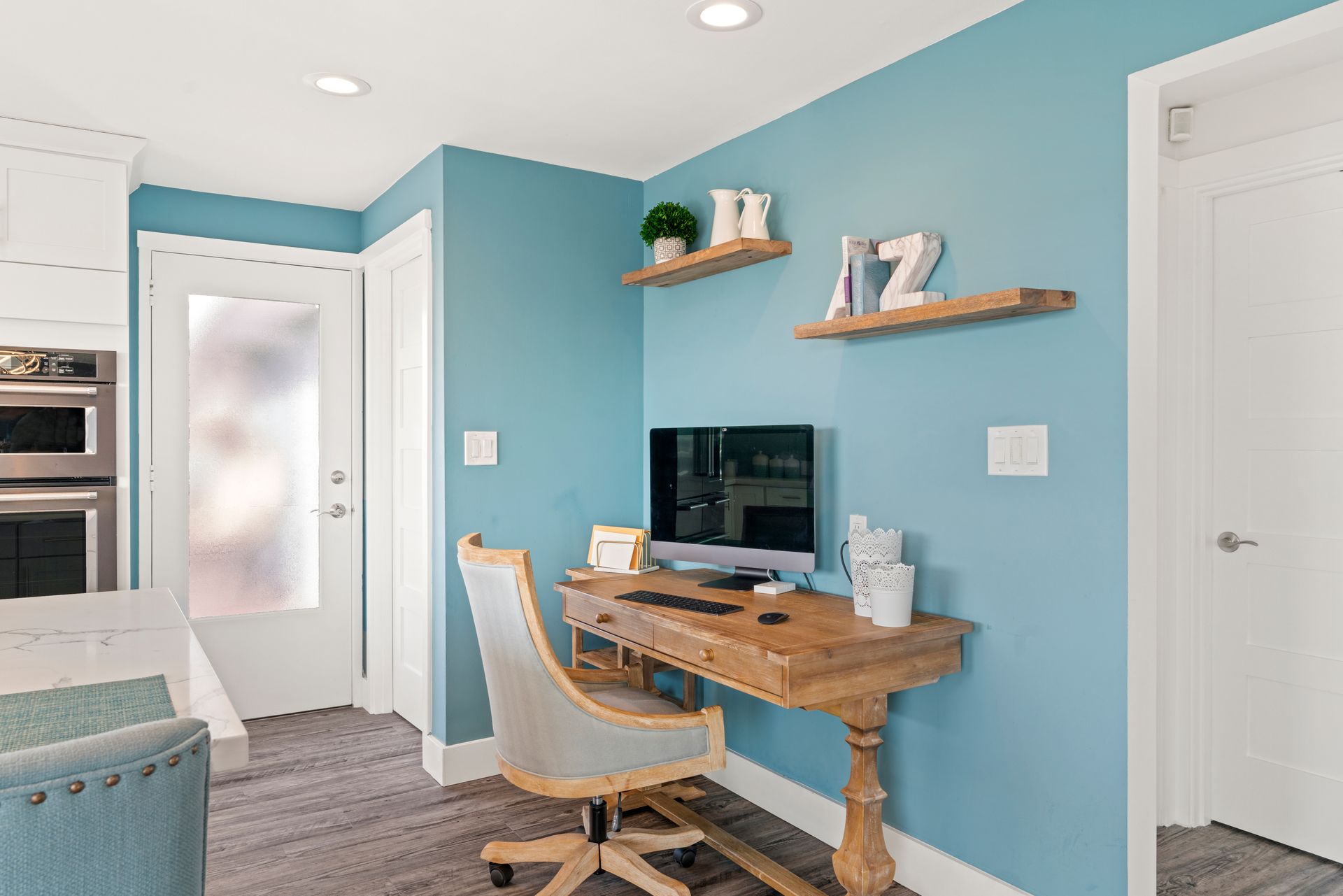 Home office nook with teal walls, wooden desk, and chair beside a bright hallway and kitchen entrance