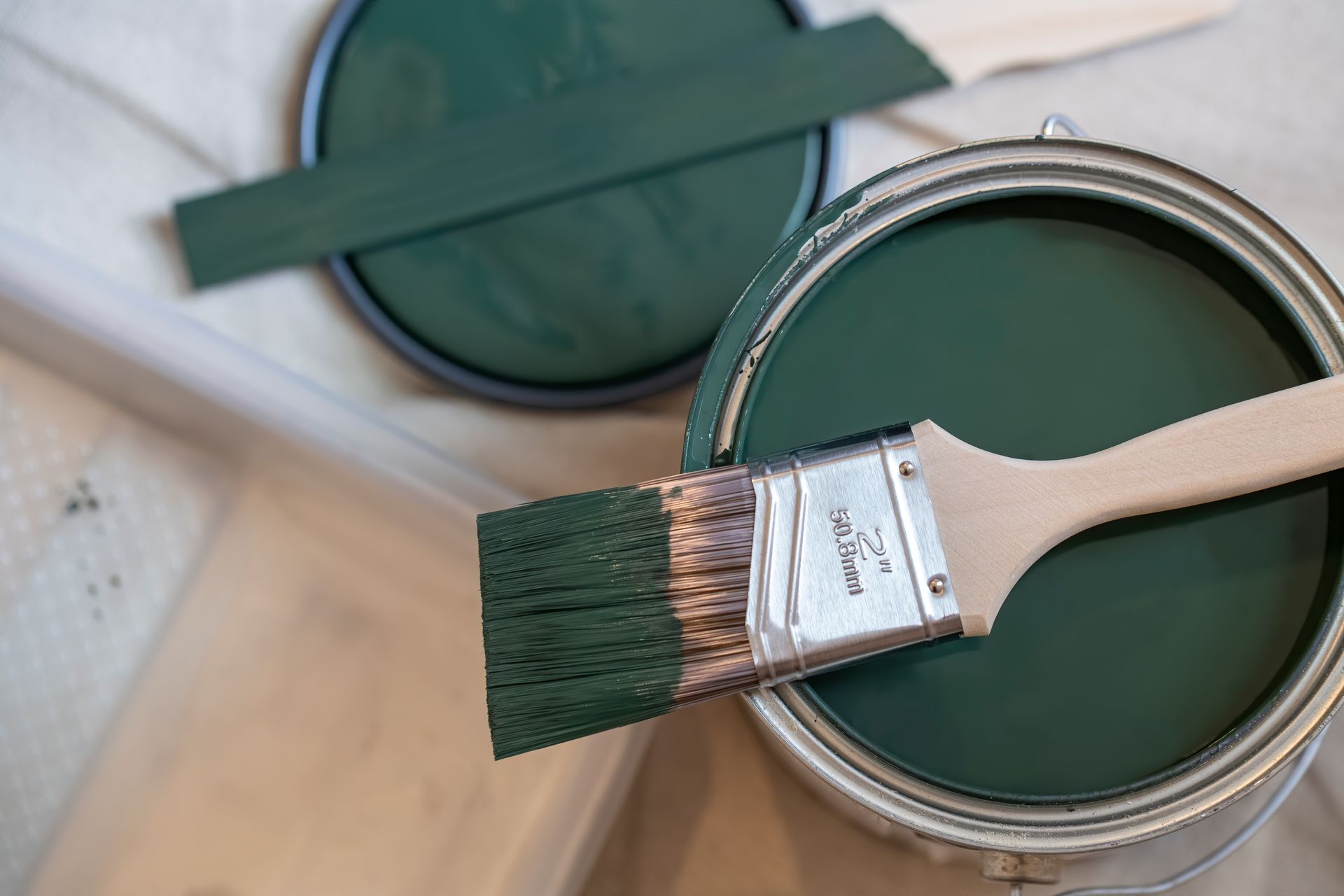 Paintbrush resting in an open can of dark green paint, with a paint lid nearby.