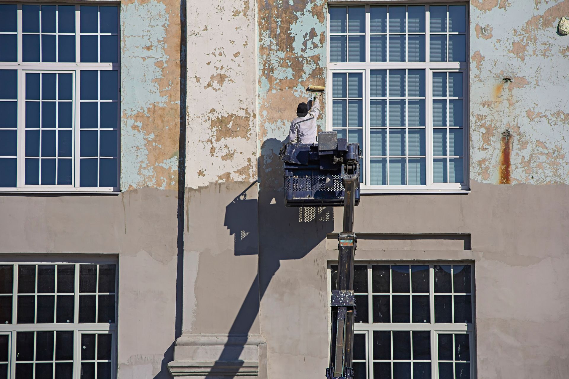 Worker on a lift painting a weathered building facade between blue windows