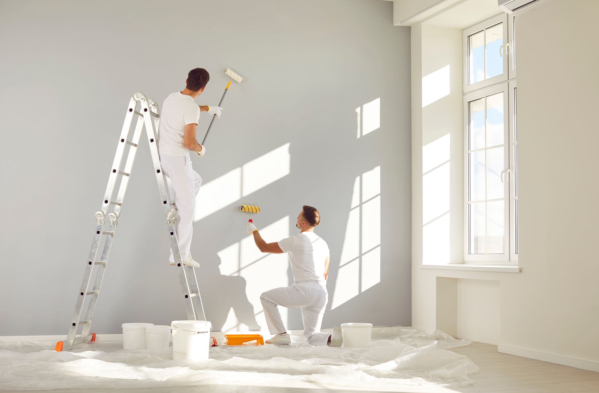 Two people painting a bright white room, one on a ladder and one kneeling with tools.