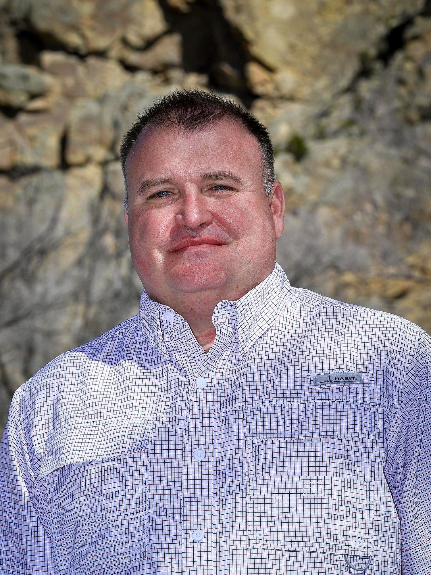 A man in a white shirt is standing in front of a rock wall.