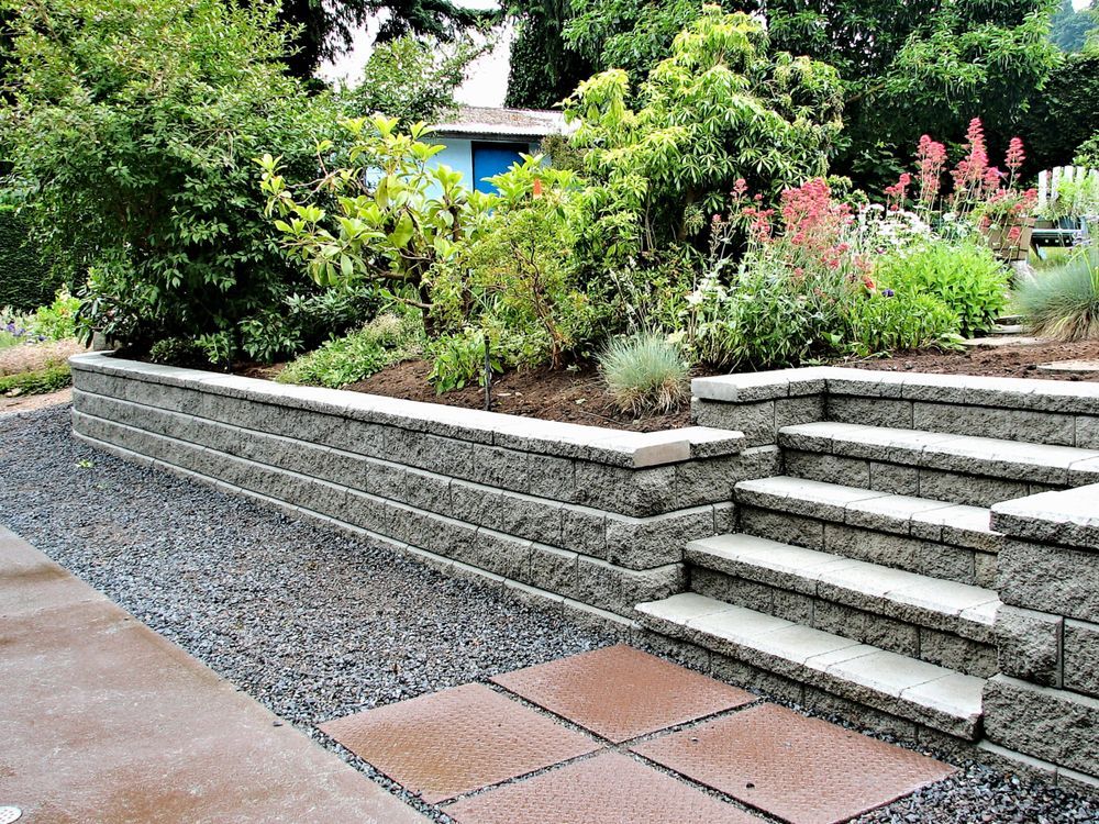 A stone retaining wall with a small set of matching stairs, set against a garden with shrubs, flowers, and gravel paths.