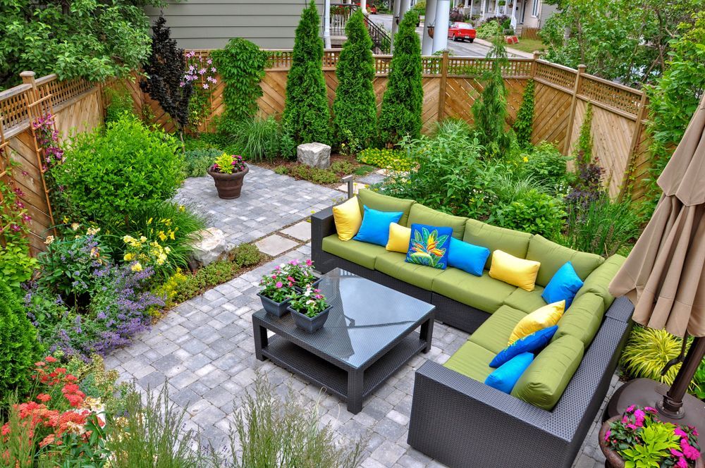 A modern backyard patio with a gray modular sofa, green cushions, blue and yellow pillows, a glass table, and garden.