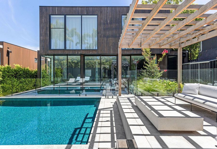Modern house with pool, shaded patio, and wooden pergola. Turquoise water, glass walls, and dark wood siding.