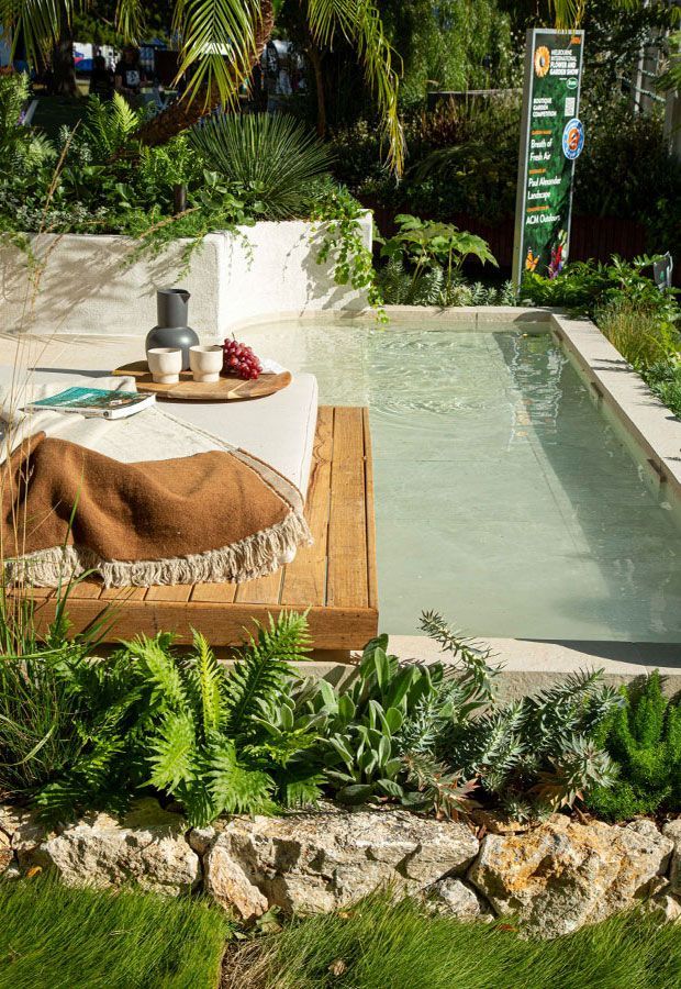 Poolside scene with a wooden deck, a blanket, and a tray of drinks. Lush greenery and stone wall surround the pool.
