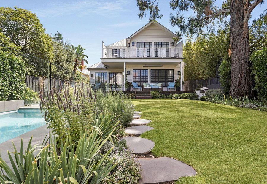 Two-story house with balcony, pool, and pathway through a grassy backyard with plants.