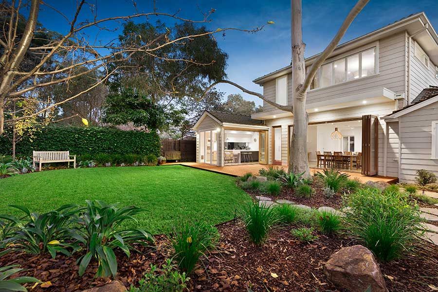 Two-story house with green lawn, wooden deck, outdoor dining, and garden beds.