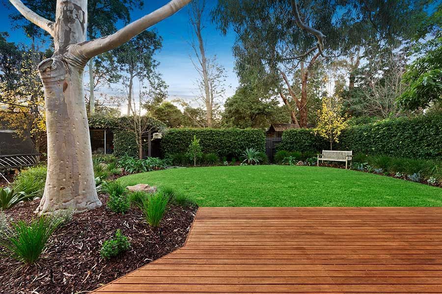 Wooden deck in front of green lawn with a tree and hedge in the background.