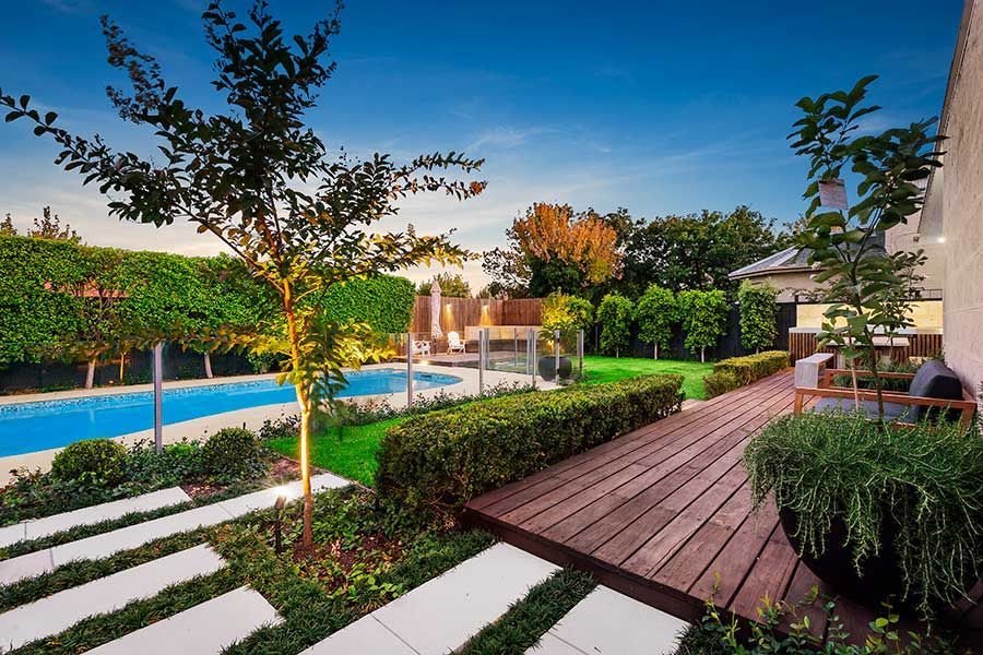 Landscaped backyard with pool, wooden deck, green hedges, trees, and blue sky.
