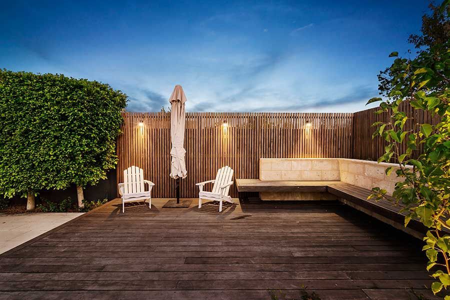 Wooden deck with white chairs, an umbrella, and a built-in bench. Wooden fence with lights. Dusk.
