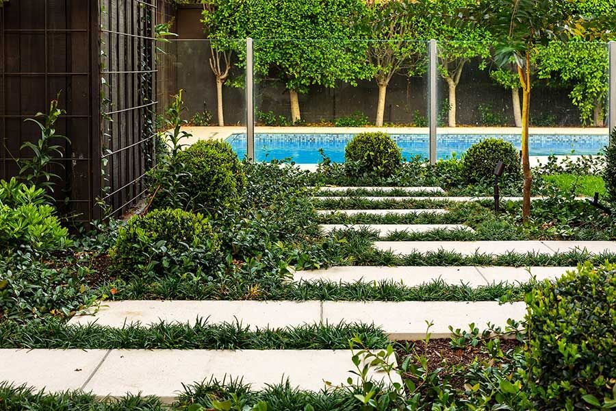 Stone pathway through a garden towards a pool. Green plants, bushes, and trees surround.