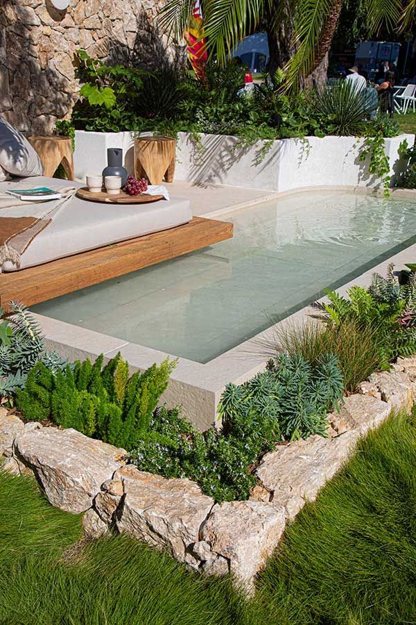 Rectangular pool with wooden deck and built-in seating surrounded by plants and a stone border.
