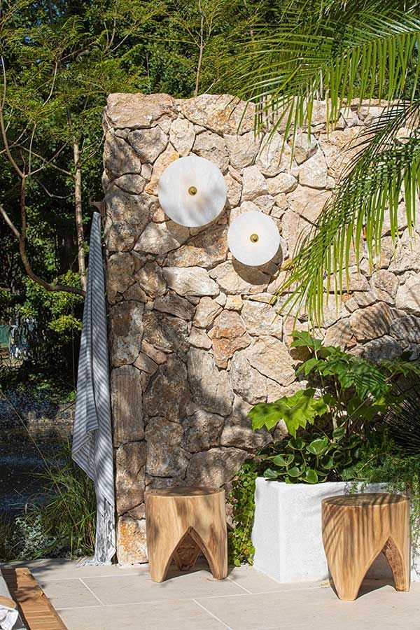 Stone wall with two round sconces, wooden stools, and plants; outdoor setting.