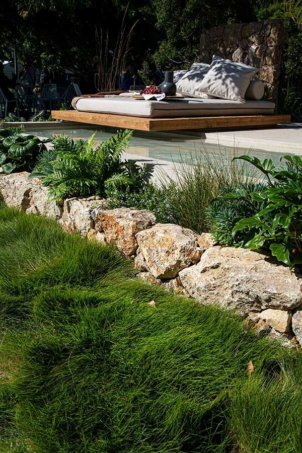 Outdoor lounge area with platform bed over a pool, next to a rock wall and green grass.