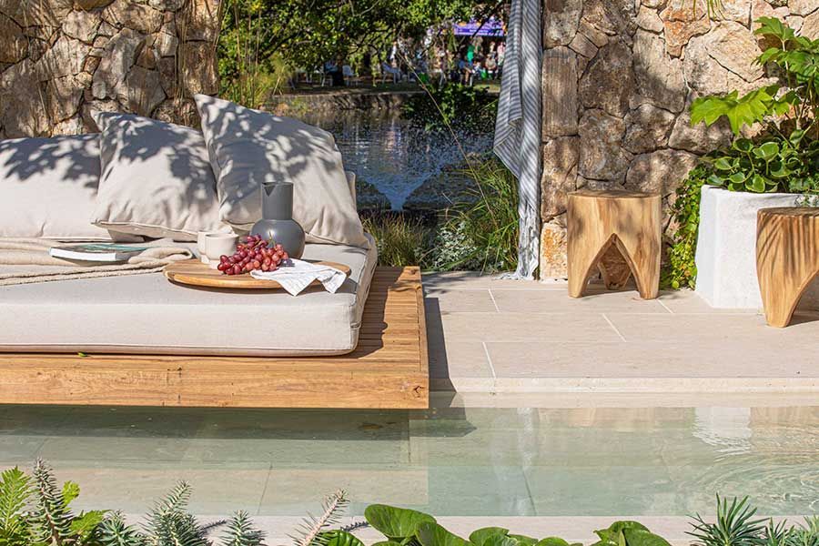 Outdoor daybed with pillows and wooden tray, set by a reflecting pool and stone wall.