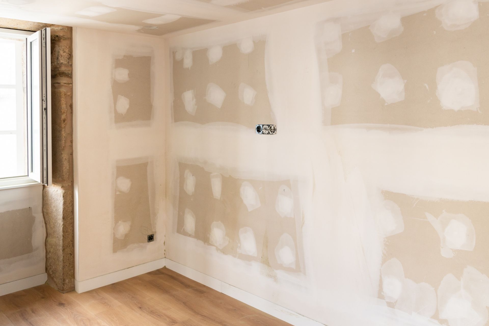 Empty room with unfinished drywall patches, white walls, wood floor, and a window on the left.