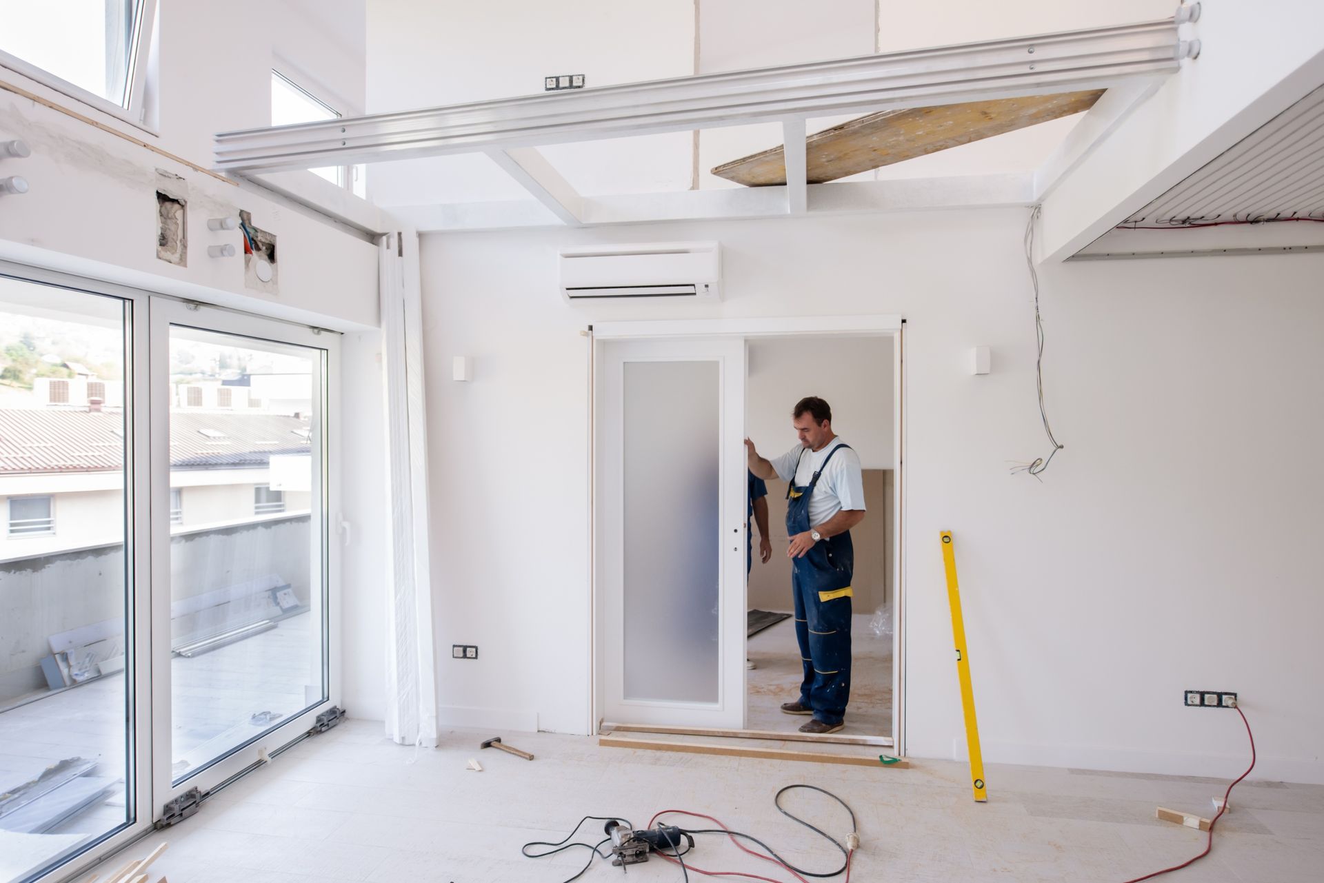 Worker installing a door in a bright, unfinished room with tools and wires on the floor
